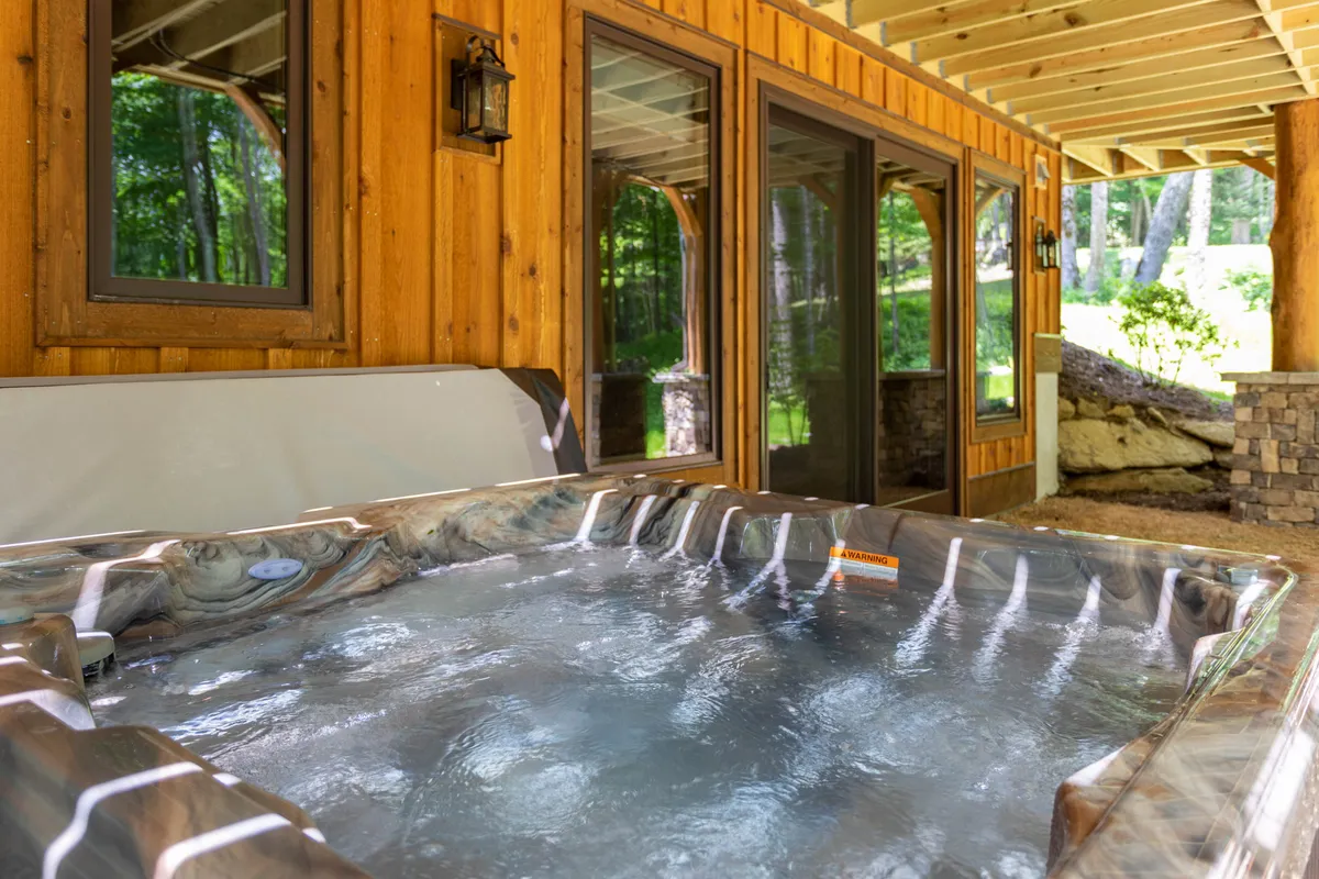 A serene outdoor hot tub nestled beside a wooden wall, with large windows reflecting the surrounding greenery. The scene conveys a sense of privacy and escape, perfect for unwinding while enjoying mountain air and natural views.