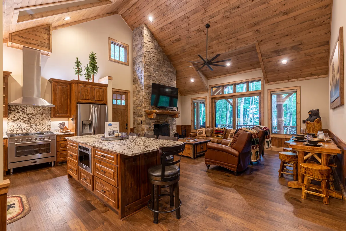 A spacious, open-concept layout unites the kitchen, dining area, and great room. The kitchen features a large granite island, while comfortable seating surrounds a stone fireplace. Expansive windows offer breathtaking forest views, emphasizing the cabin’s seamless connection to nature and its capacity for hosting groups.