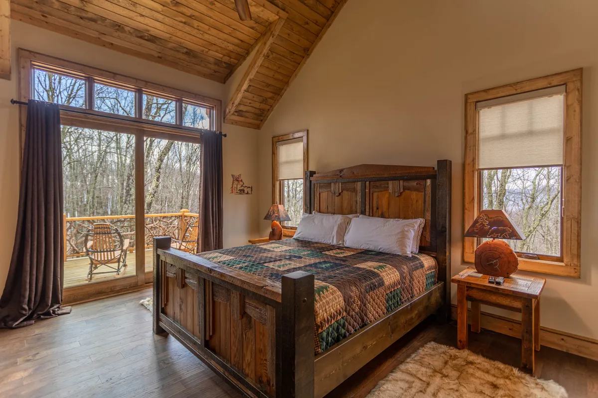 A tranquil master suite with a wooden bed, large windows letting in natural light, decorative lamps on the nightstands, and a cozy rug. The arrangement conveys comfort and privacy, while the view through the windows connects the space to the surrounding nature.