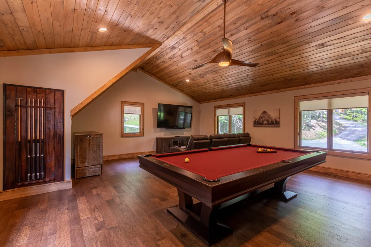 An inviting game room featuring a sleek pool table at the center, cozy sofa seating nearby, a television set along the wall, and large windows letting in natural light. The wood-paneled ceiling and relaxed furnishings create a welcoming, playful vibe ideal for gatherings.
