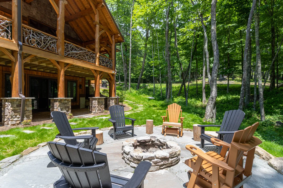A picturesque exterior gathering area featuring a stone fire pit encircled by Adirondack chairs, with lush greenery and wooden beams of the house in the background. The mood is cozy and communal, ideal for storytelling and marshmallow roasting.
