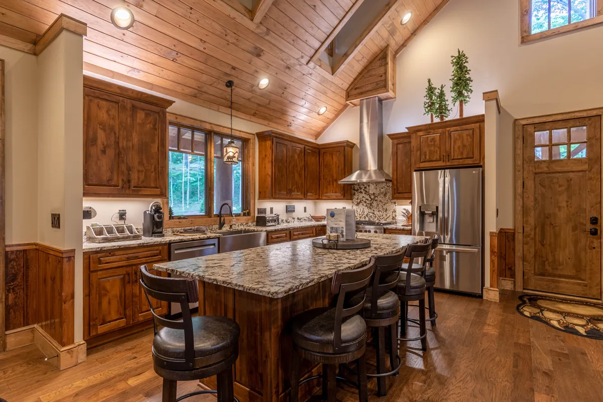 A chef’s kitchen with gleaming granite countertops, rich wooden cabinetry, stainless steel appliances, and a central island with bar seating. Large windows bring in natural light, creating an inviting space for cooking, casual breakfasts, or gathering while meals are being prepared.