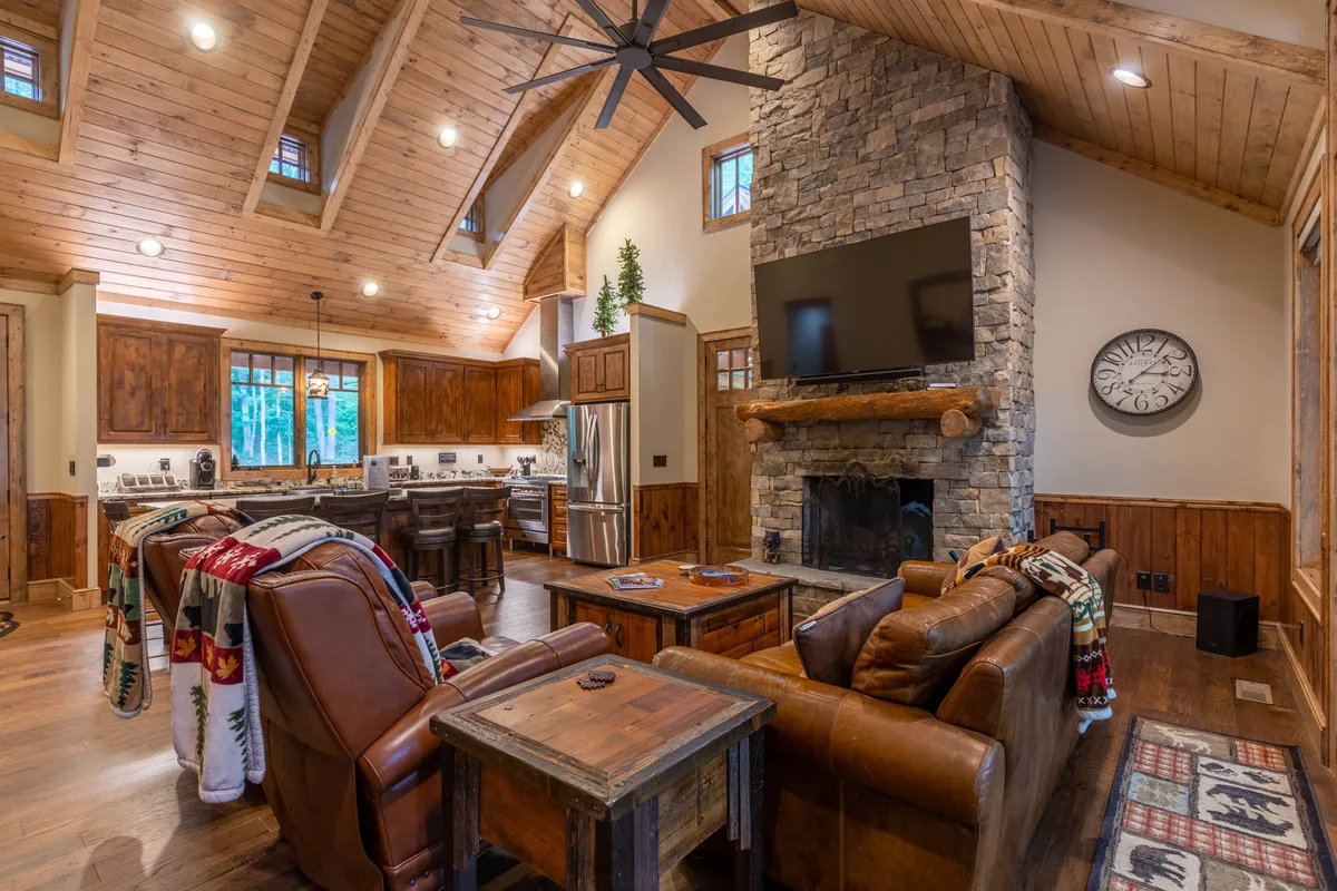 A warm, welcoming great room featuring a grand stone fireplace as its centerpiece, surrounded by comfortable leather sofas. Large windows flood the space with natural light and frame panoramic woodland views, while the open layout allows you to glimpse the adjacent kitchen and dining area. Subtle decor and inviting seating create an ideal spot for gatherings, movie nights, or relaxing by the fire.