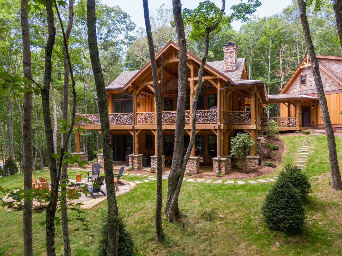 A view of a wooden balcony with an intricate railing, a stone pathway leading to a fire pit area with chairs, and lush greenery surrounding the property. This setup highlights areas for outdoor gatherings, relaxation, and connecting with nature, all within steps of the cabin.