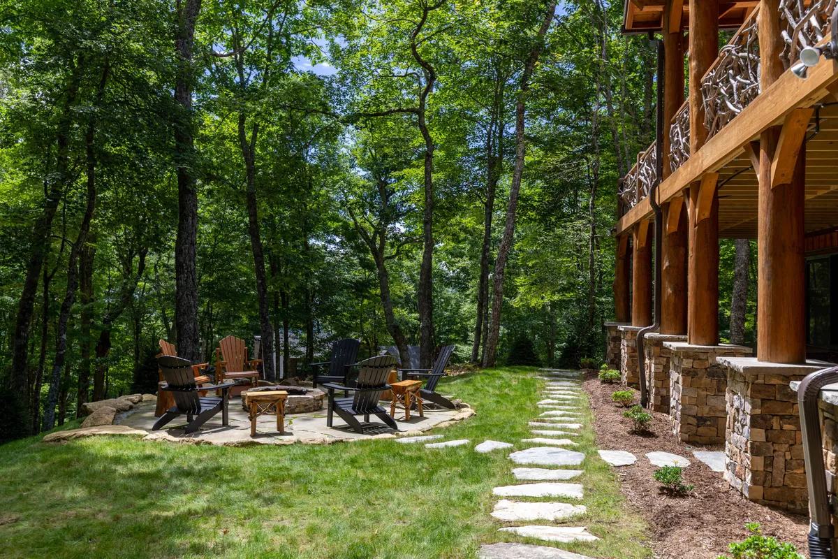 A cozy outdoor fire pit encircled by Adirondack chairs rests beside a wooden structure and stone pathway, surrounded by trees. The setting invites guests to gather for storytelling, warmth, and stargazing in a peaceful woodland environment.