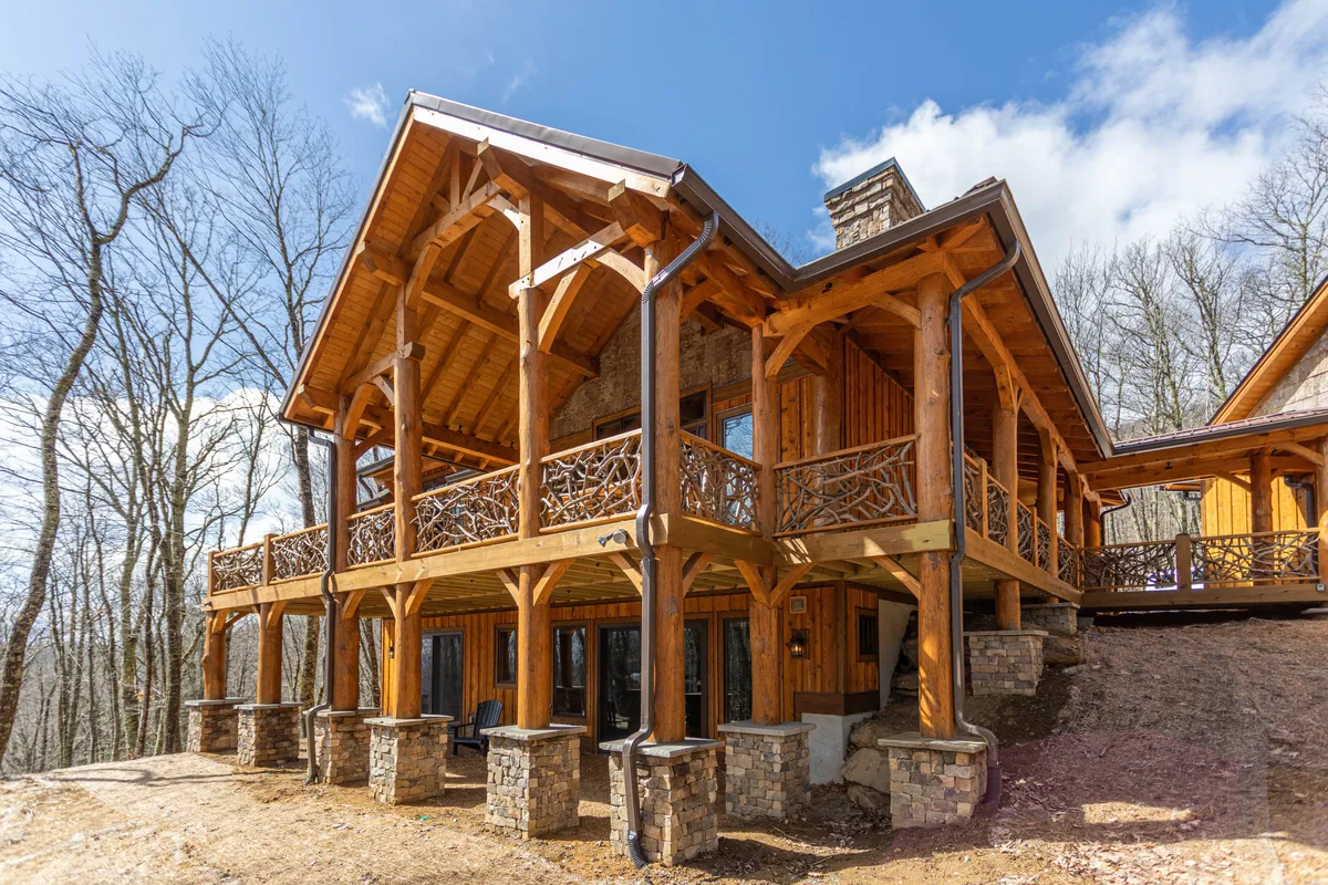 A cozy yet grand exterior of a luxury mountain cabin, featuring wooden beams, stone columns, a balcony railing, and large windows. The setting is perfect for outdoor gatherings, BBQs, or simply relaxing while soaking in sunset views.