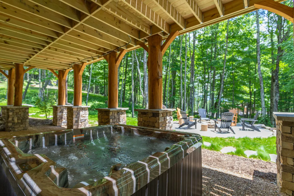 This exterior image features a hot tub set against a forested backdrop, with wooden pillars, stone columns, and adirondack chairs creating a welcoming and tranquil setting. The space feels private yet open to nature, ideal for relaxation or group gatherings under the stars.