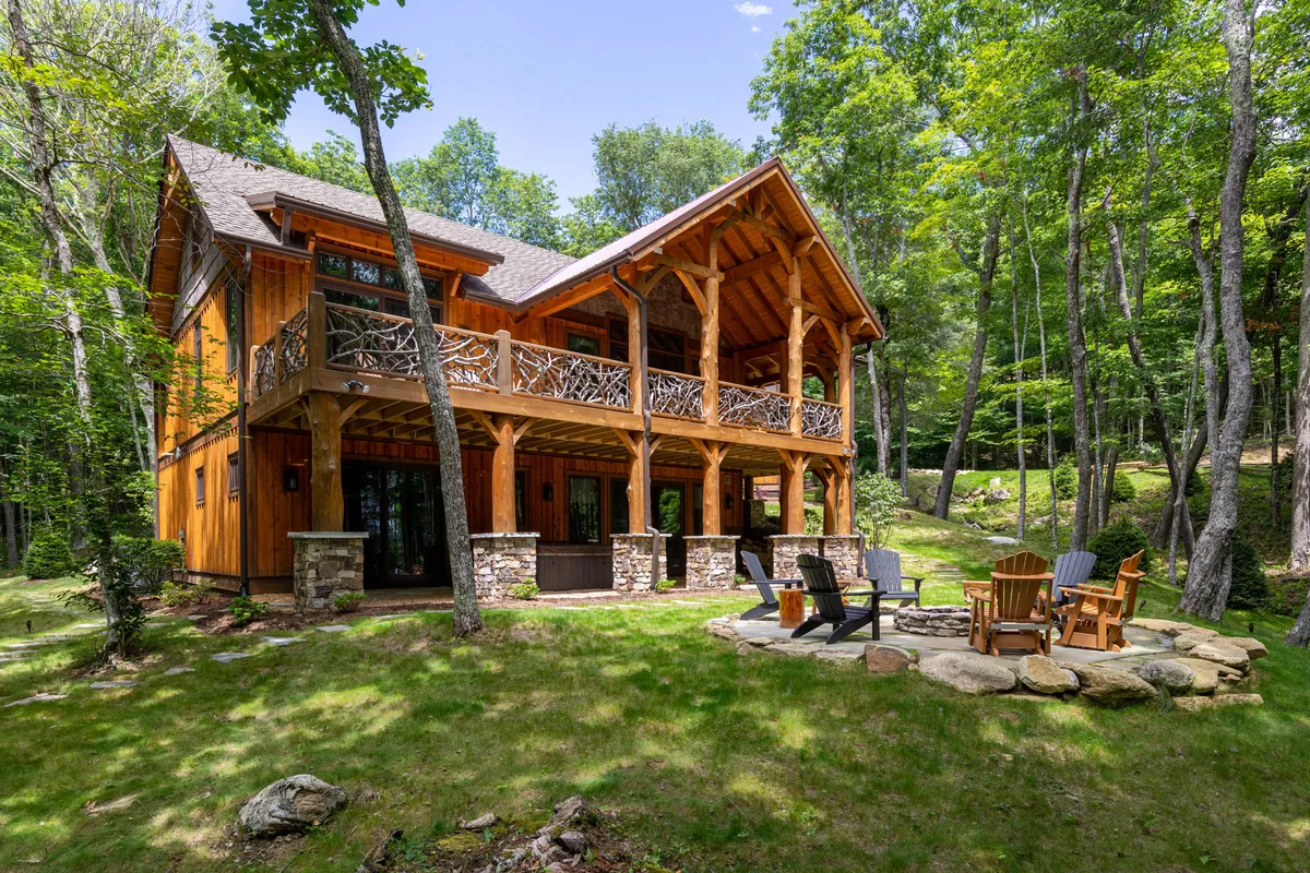 A tranquil outdoor area with a wooden balcony, stone fire pit, surrounding trees, and expansive windows. The atmosphere is inviting for stargazing, morning coffee, evening bonfires, and group gatherings, blending natural beauty with cabin comfort.