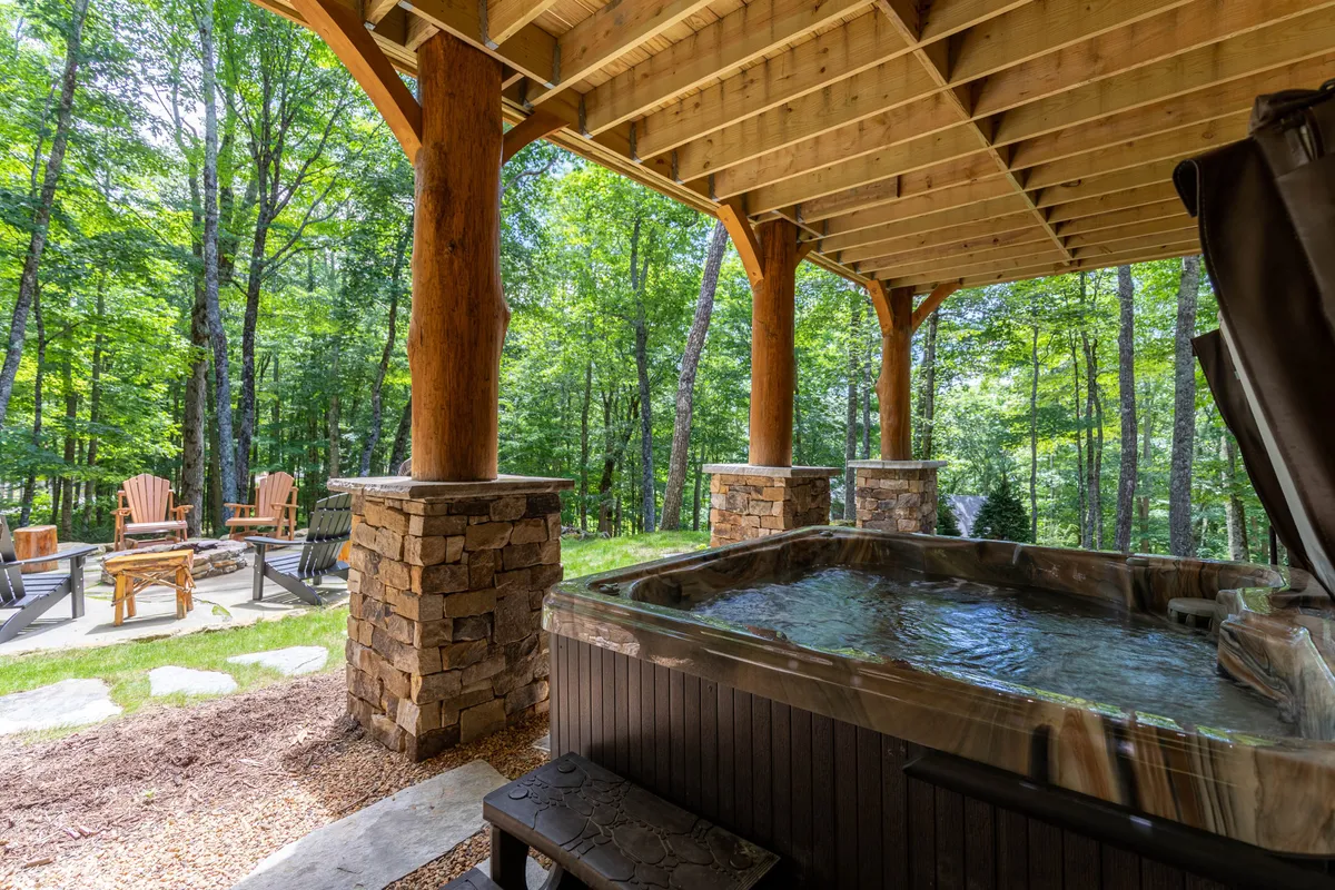 The outdoor hot tub sits under a covered area with sturdy wooden pillars and stone columns, surrounded by Adirondack chairs and lush greenery. The scene evokes peace and privacy, ideal for soaking after a day of adventure and gazing at the sunset or stars.