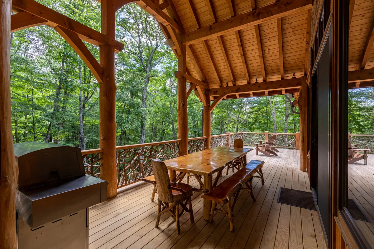 A spacious wooden dining table and grill set on a deck, surrounded by tall trees and Adirondack chairs. This inviting outdoor space is ideal for shared meals, morning coffee, and group gatherings, blending seamlessly with the natural environment.